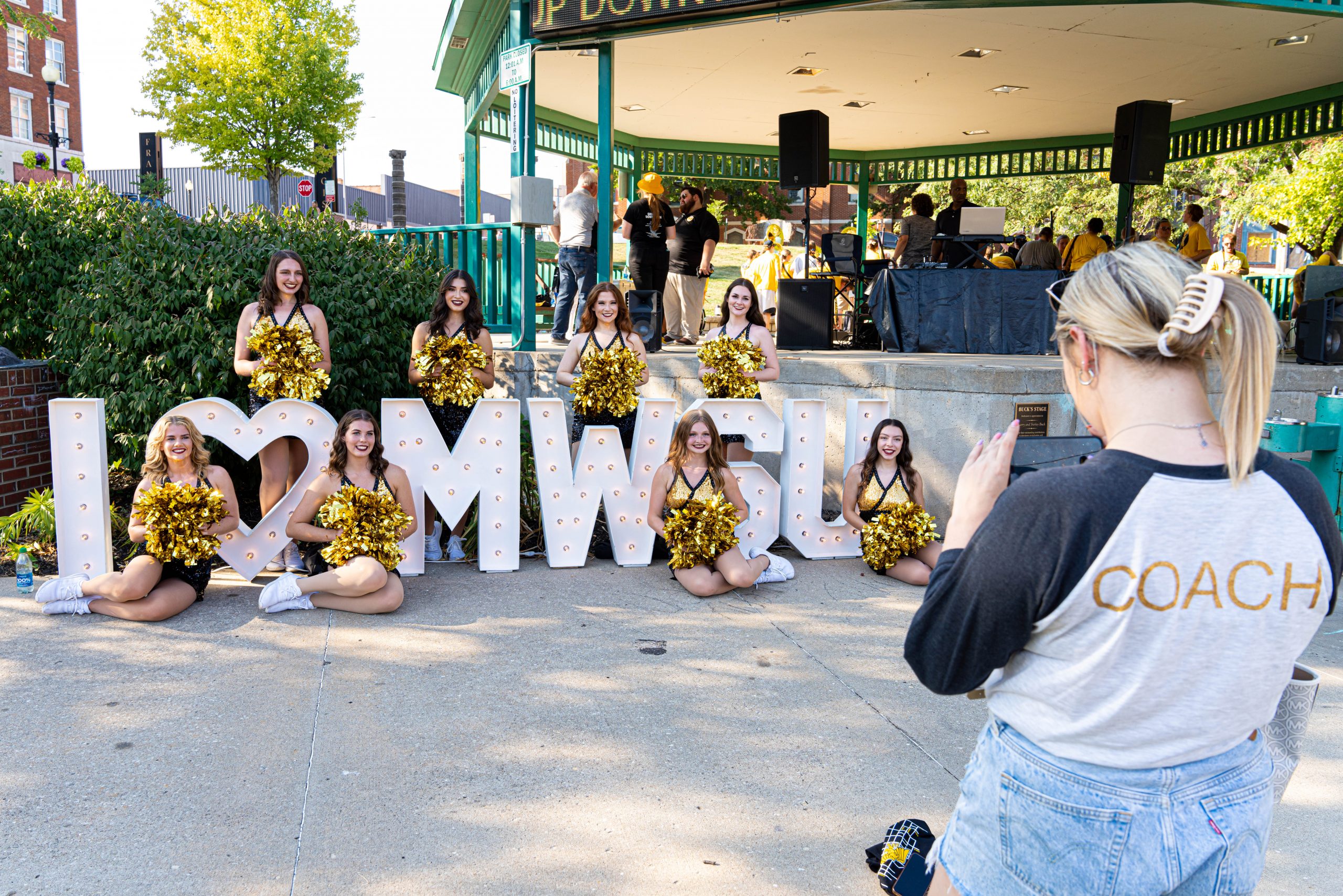 individual with coach on their shirt taking a photo of missouri western cheer leaders posing with a sign that reads i heart mwsu